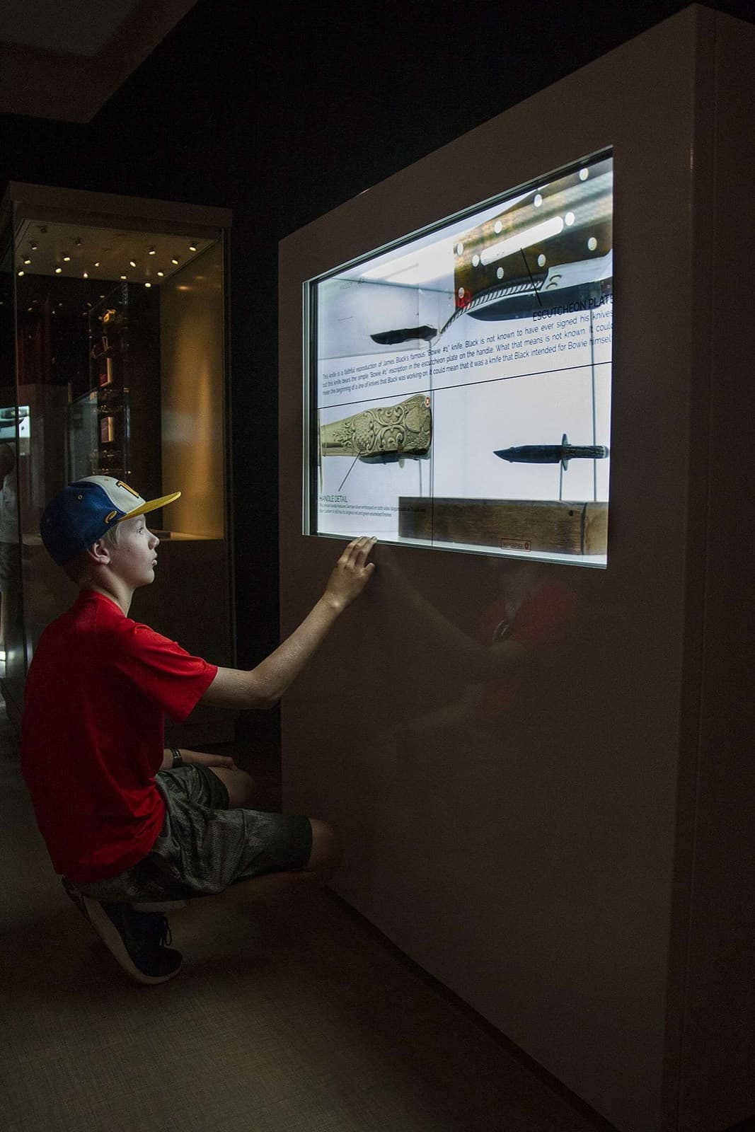 A child kneeling and touching a wall-mounted translucent touch screen showing knife artifacts.